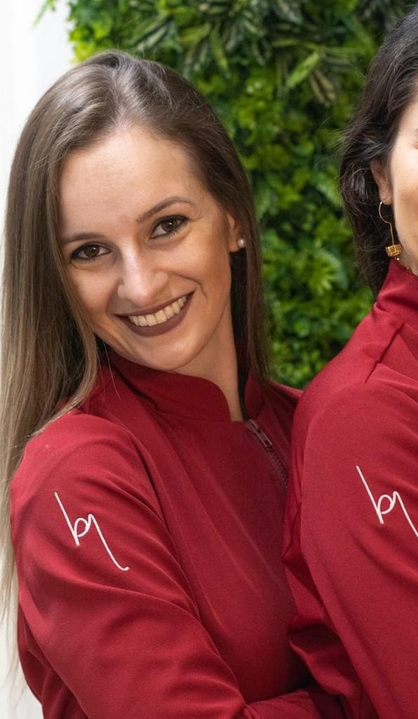Dra. Camila Cauduro, de jaleco framboesa, sorrindo em frente ao jardim vertical