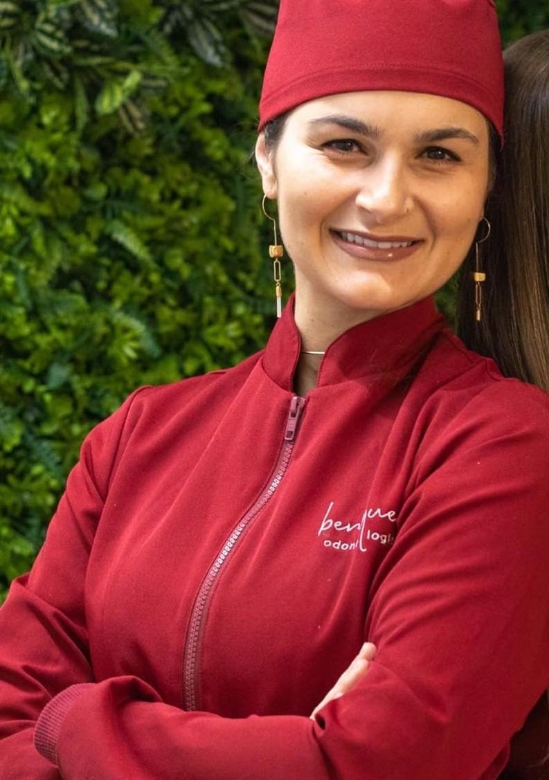 Dra. Caroline Ló Guarnieri, de jaleco e touca framboesa, sorrindo em frente ao jardim vertical